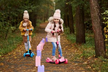 Two girls having fun on scooters in a leafy autumn park trail.